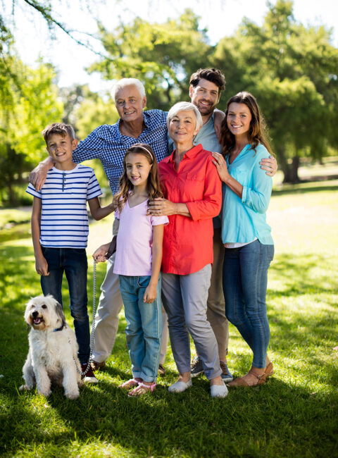Three generations of a smiling family—grandparents, parents, and two children—stand together with arms around each other in a sunny park, with a fluffy white dog sitting in front on the grass.