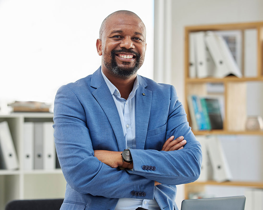 A smiling man in a blue suit jacket stands with his arms crossed in an office setting, with shelves of files and books in the background.