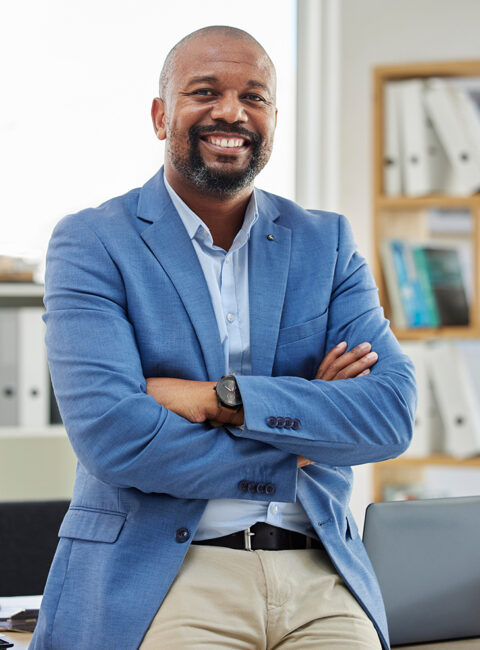 A smiling man in a blue blazer and beige pants sits on a desk with his arms crossed in a modern office, with shelves of binders and a laptop visible in the background.