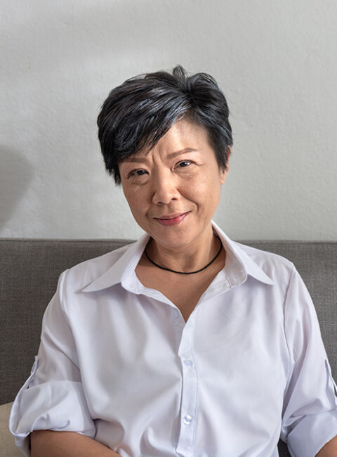 A woman with short dark hair, wearing a white button-up shirt and a black necklace, sits on a gray couch against a plain light-colored wall, smiling gently at the camera.