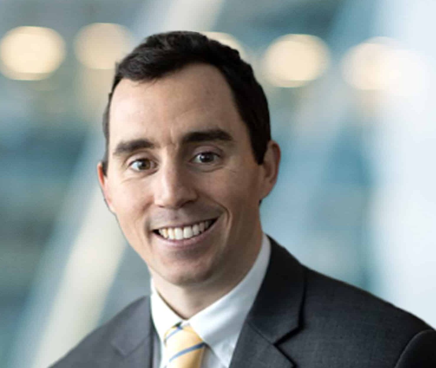 A man in a dark suit, white shirt, and yellow-striped tie smiles at the camera with a blurred office background.