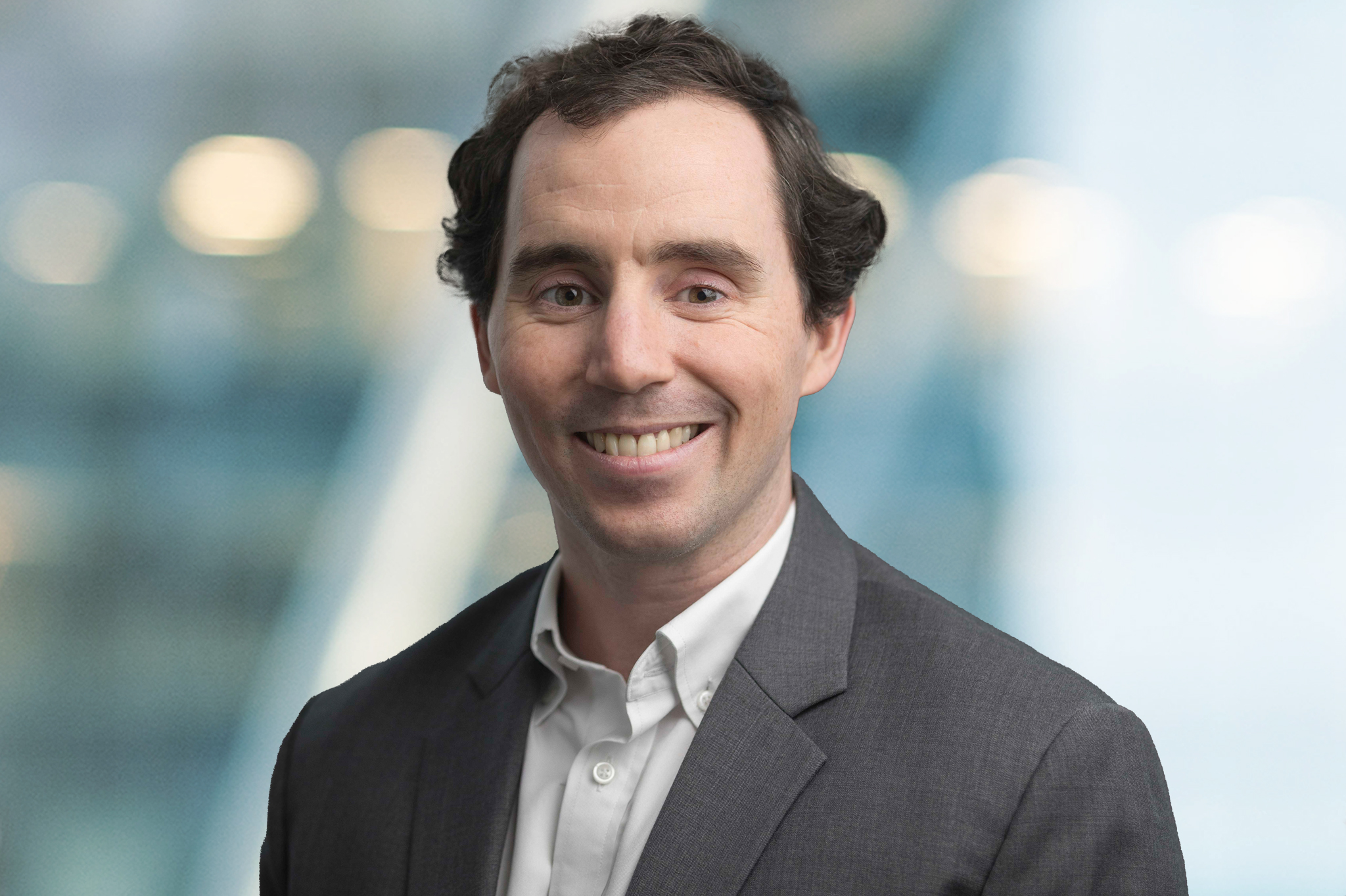 A man with short, curly brown hair wearing a grey suit jacket and white shirt smiles at the camera. The background is softly blurred with blue and yellow tones.