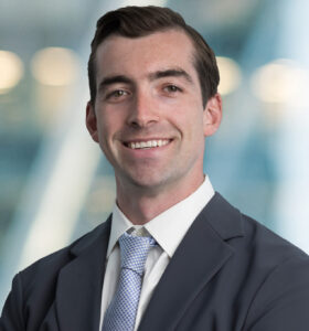 A young man in a gray suit and light blue tie smiles confidently with arms crossed, standing in front of a bright, blurred office background.