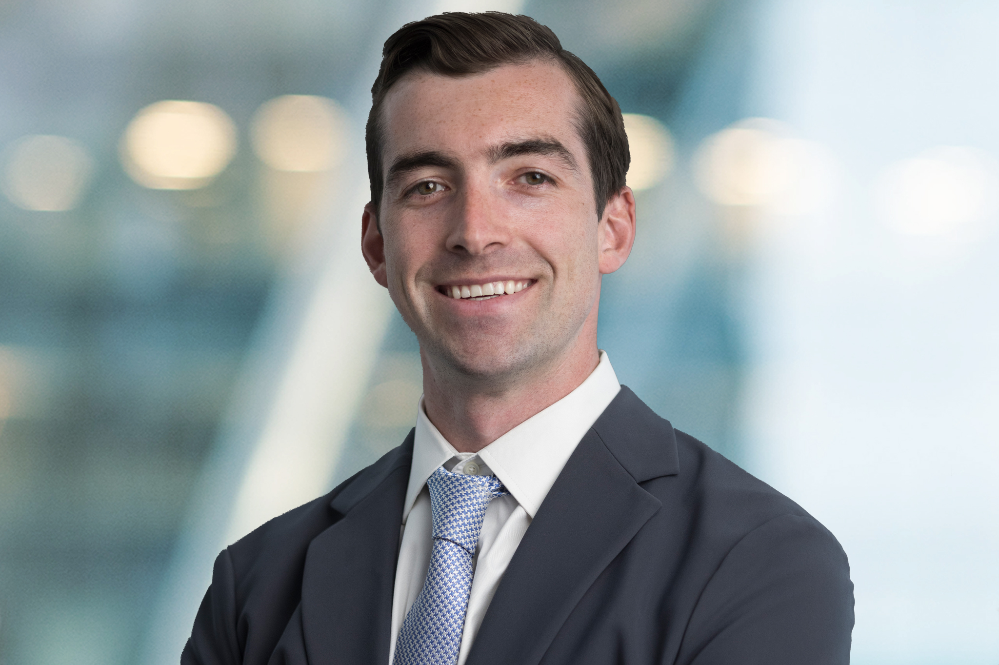 A young man in a gray suit and light blue tie smiles confidently with arms crossed, standing in front of a bright, blurred office background.