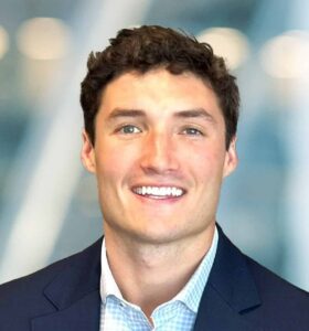 A young man with short, curly brown hair smiles at the camera. He is wearing a navy blue blazer over a light blue checkered shirt, and the background is softly blurred with blue and white tones.