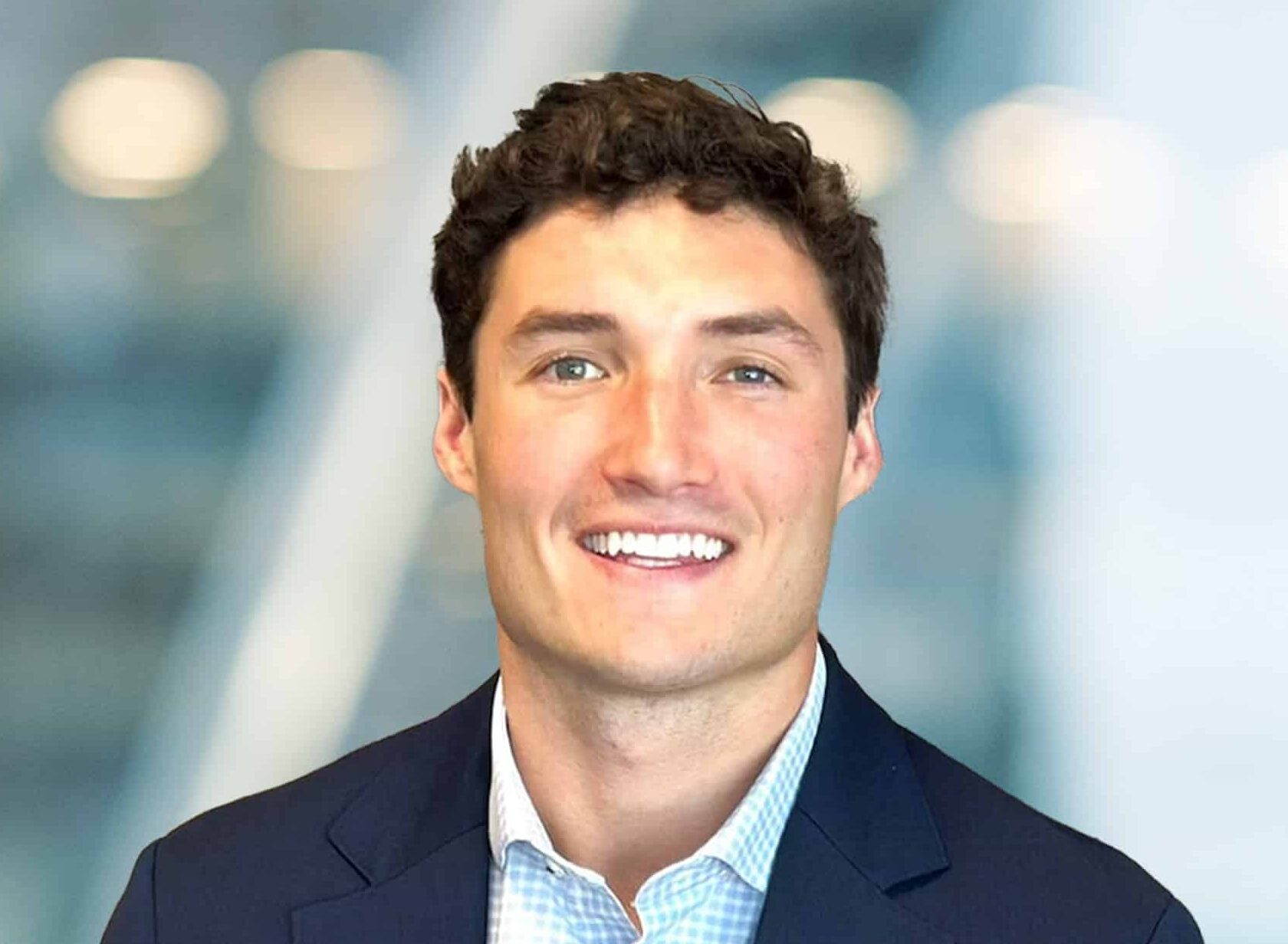 A young man with short, curly brown hair smiles at the camera. He is wearing a navy blue blazer over a light blue checkered shirt, and the background is softly blurred with blue and white tones.