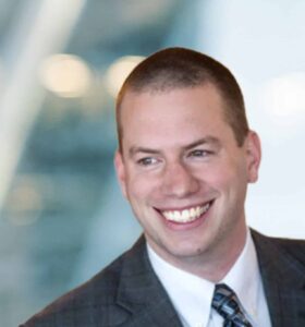 A man wearing a plaid suit, white shirt, and patterned tie smiles broadly while looking to the side; the background is blurred with soft, bright lighting.