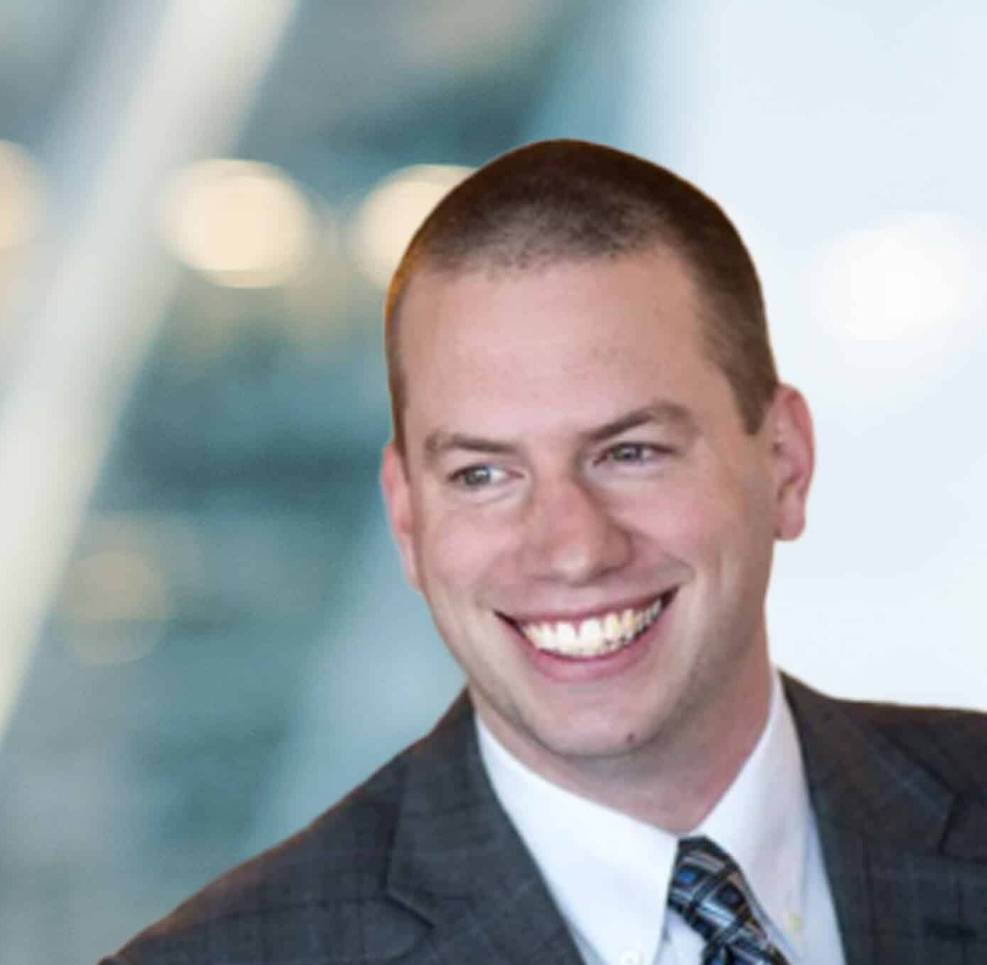 A man wearing a plaid suit, white shirt, and patterned tie smiles broadly while looking to the side; the background is blurred with soft, bright lighting.