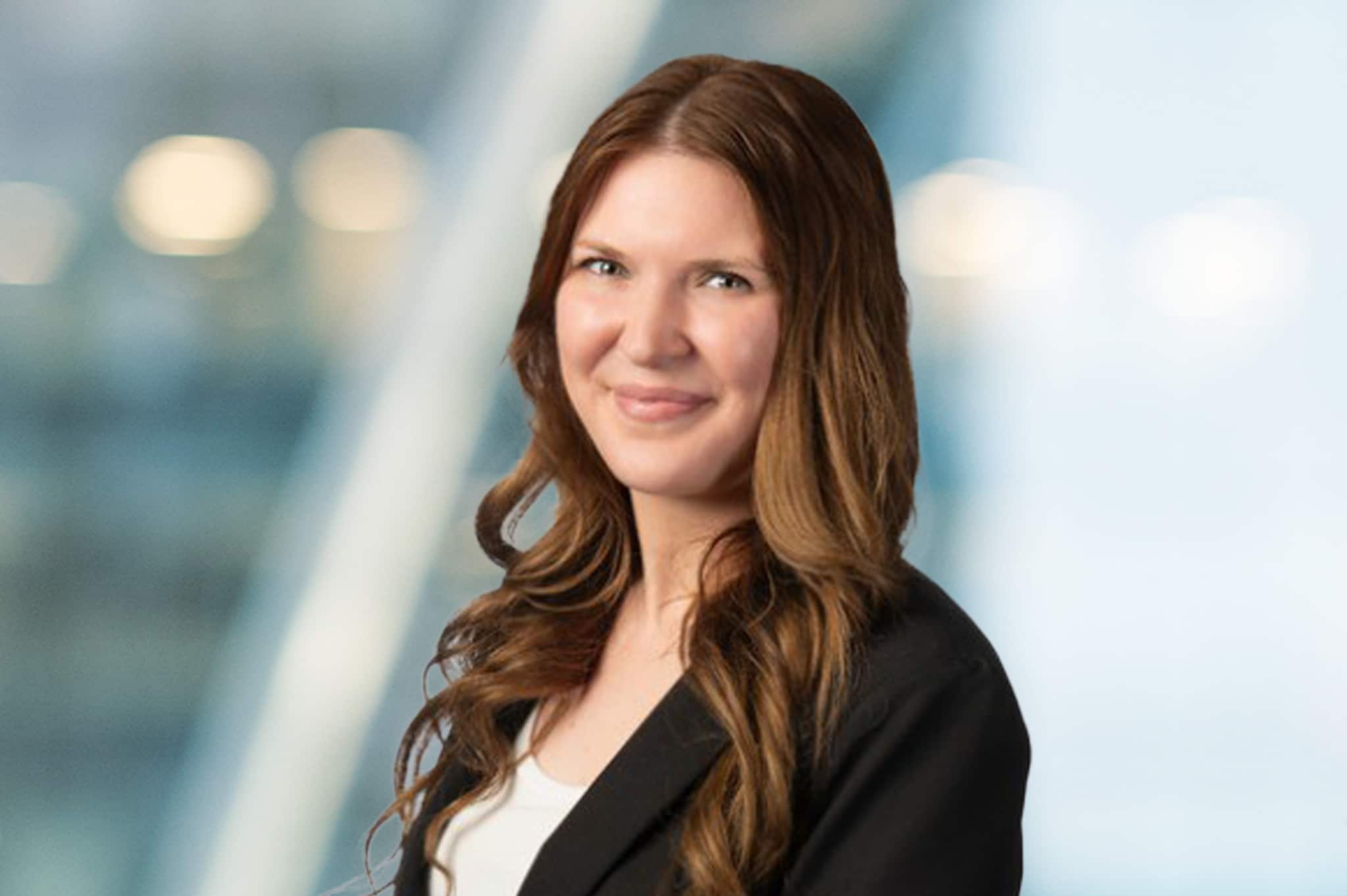 A woman with long, wavy brown hair wearing a black blazer and white top smiles in front of a blurred, modern indoor background.