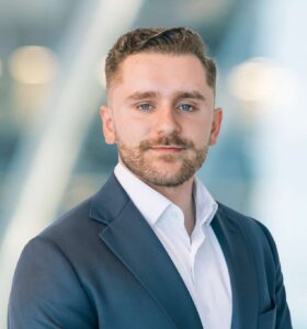 A man with short brown hair and a trimmed beard wears a dark suit jacket over a white shirt, posing in front of a blurred, modern office background.