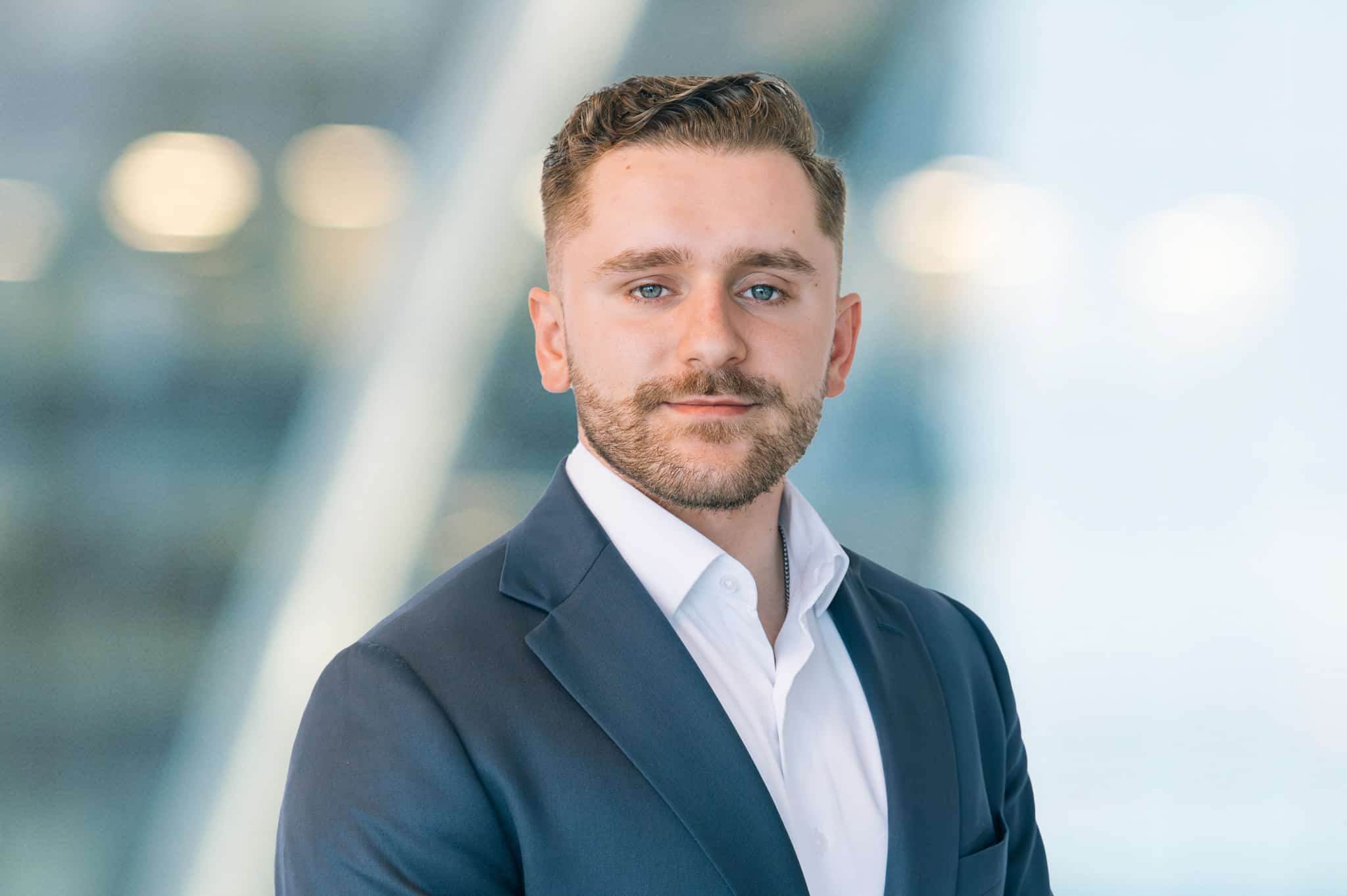 A man with short brown hair and a trimmed beard wears a dark suit jacket over a white shirt, posing in front of a blurred, modern office background.
