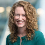A woman with curly, shoulder-length blonde hair smiles at the camera. She is wearing a textured green top and stands in front of a blurred background with soft lights.