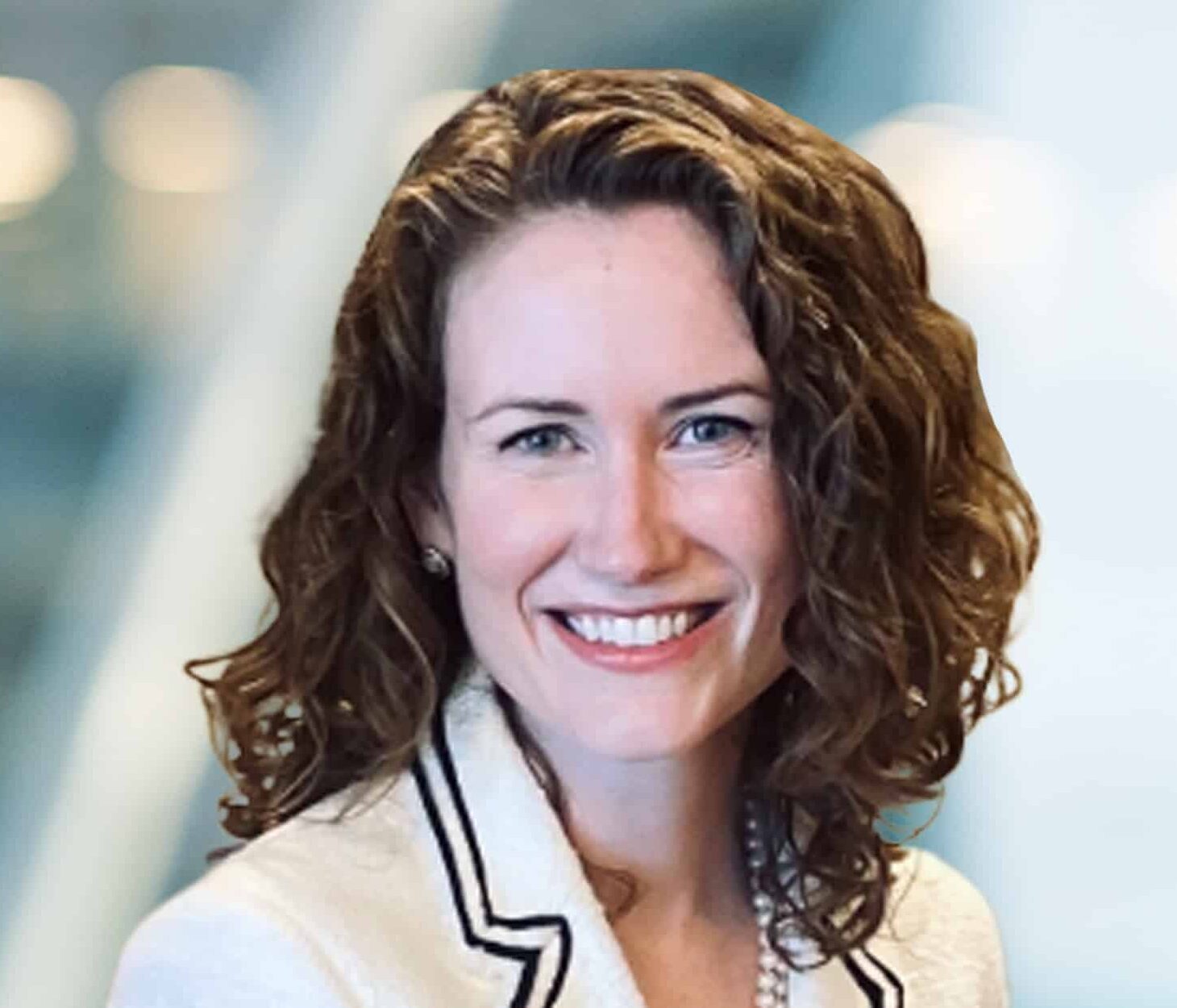 A woman with curly brown hair, wearing a white blazer with dark trim and a pearl necklace, smiles at the camera against a blurred, light-colored background.