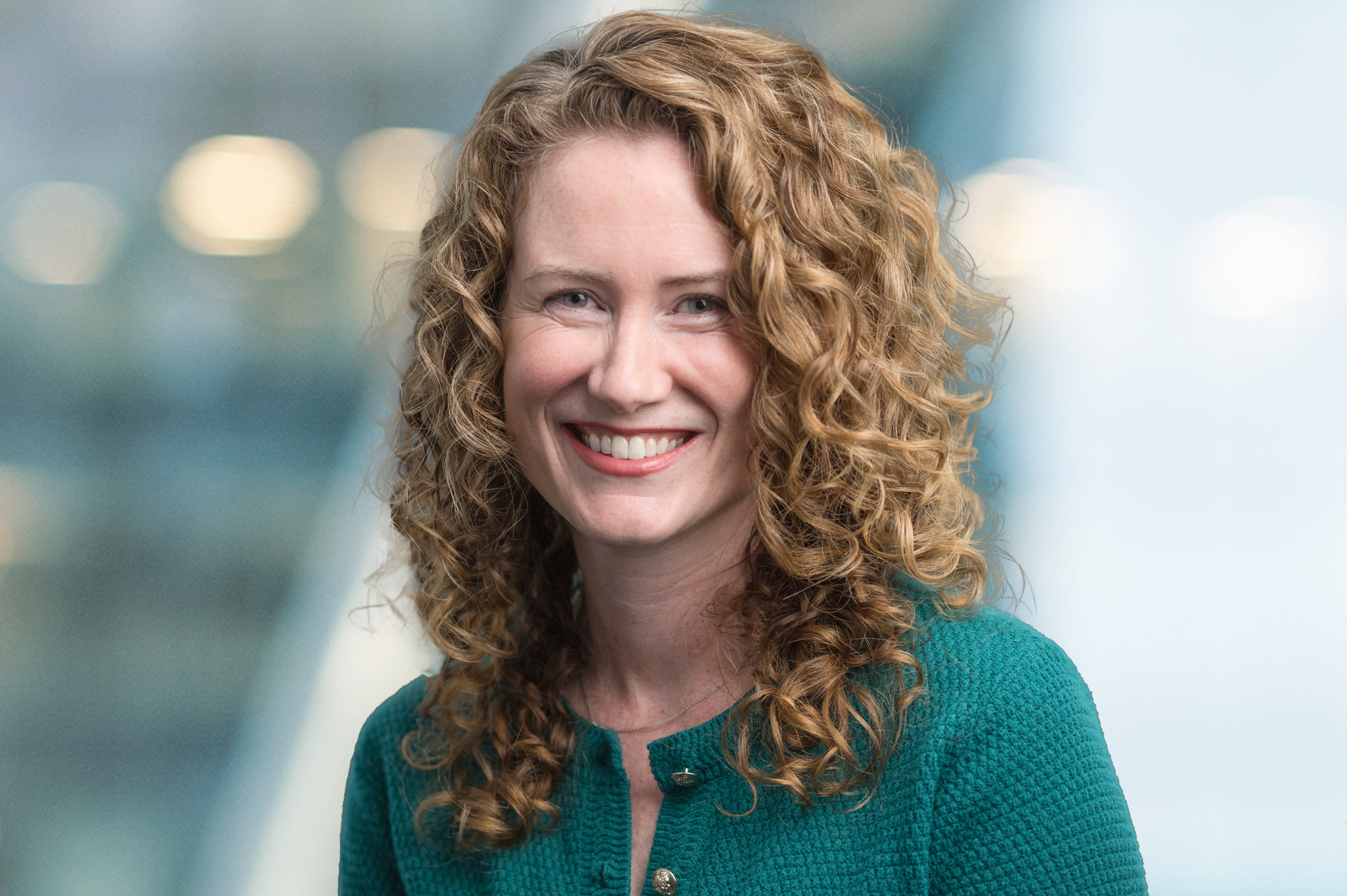 A woman with curly, shoulder-length blonde hair smiles at the camera. She is wearing a textured green top and stands in front of a blurred background with soft lights.