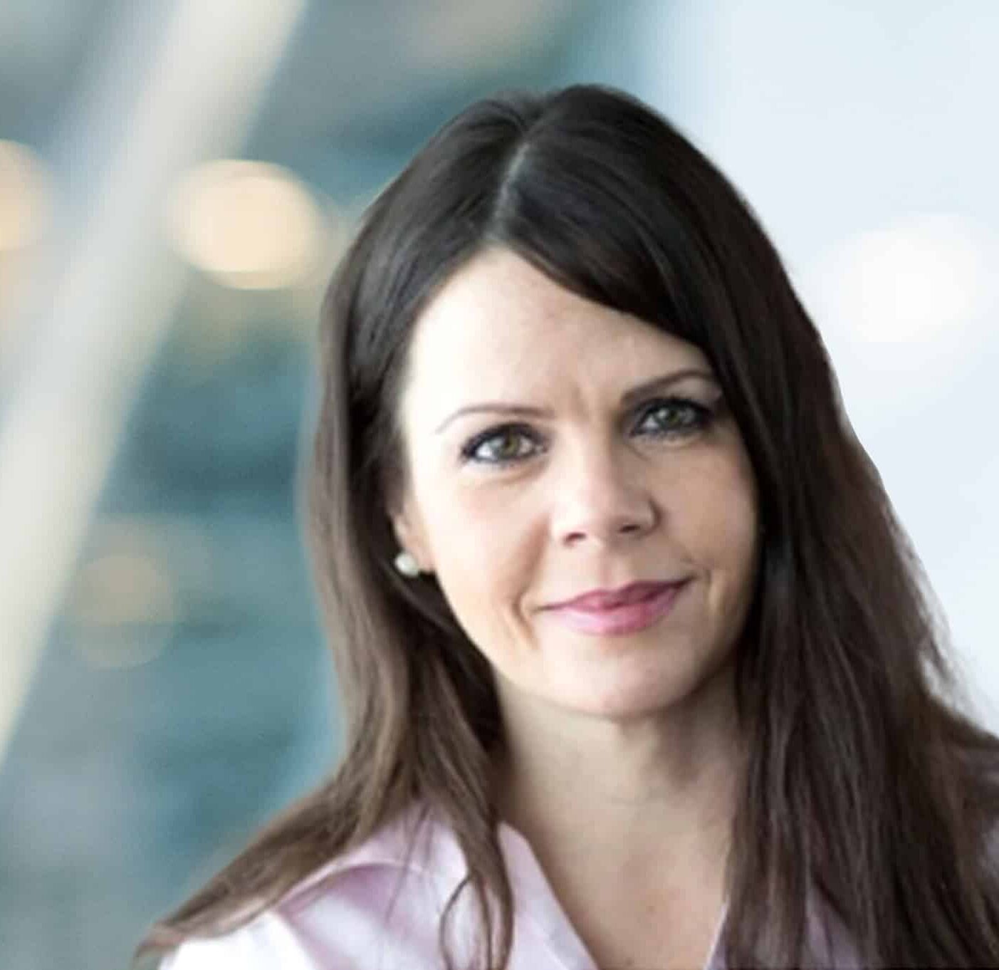 A woman with long brown hair and light skin, wearing a light pink collared shirt, is smiling softly at the camera. The background is blurred with blue and light tones.