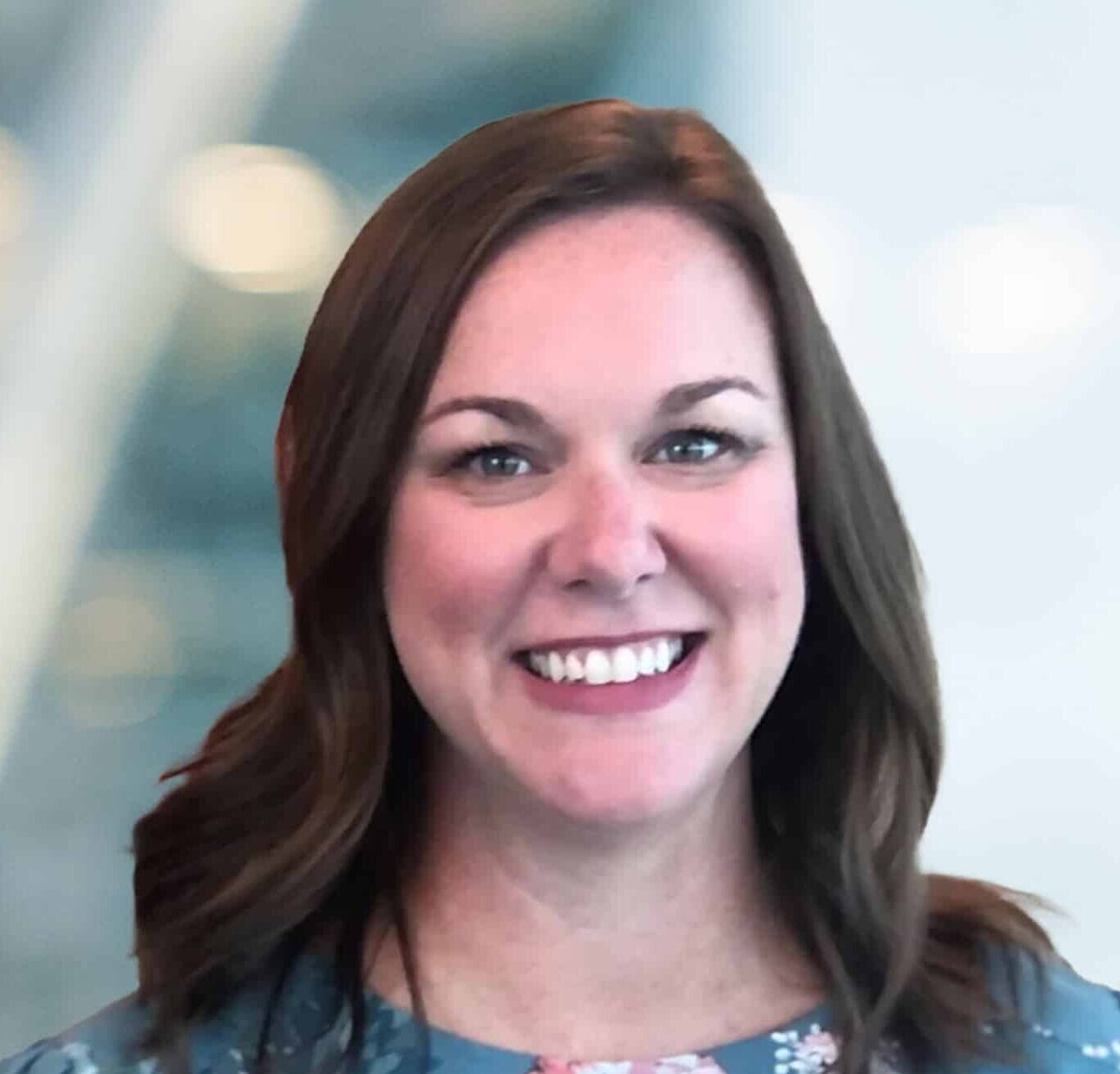 A woman with shoulder-length brown hair, wearing a floral top, smiles at the camera. The background is softly blurred with cool tones and circular light spots.