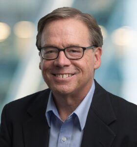 A middle-aged man with short light brown hair, glasses, and a blue collared shirt under a dark blazer, smiling in front of a blurred background with soft lights.