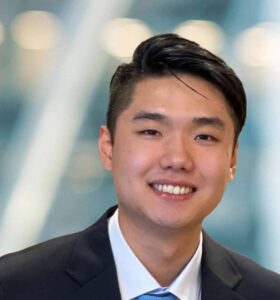 A young man wearing a dark suit and blue tie is smiling at the camera, with a blurred background featuring soft lights and diagonal lines.