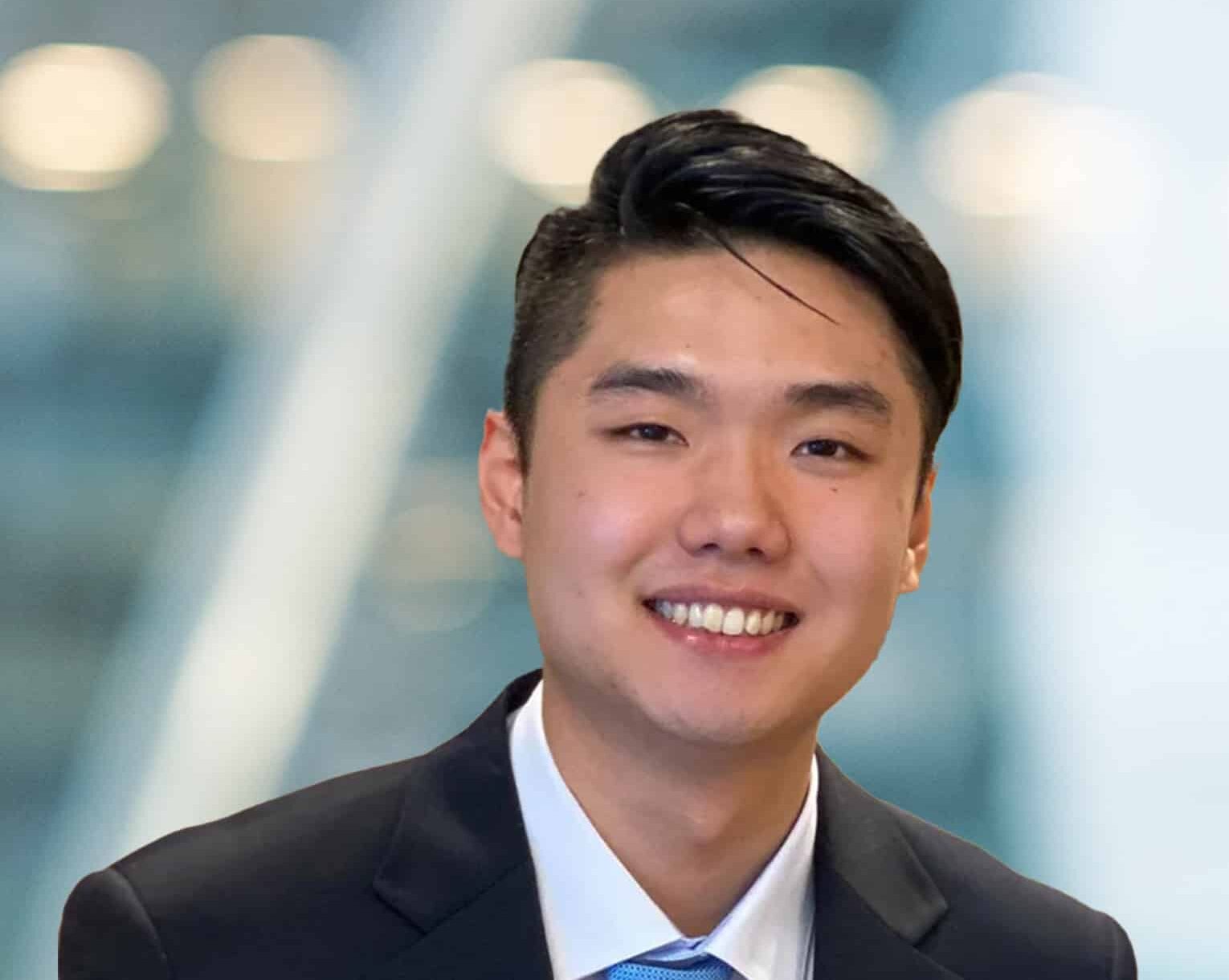 A young man wearing a dark suit and blue tie is smiling at the camera, with a blurred background featuring soft lights and diagonal lines.