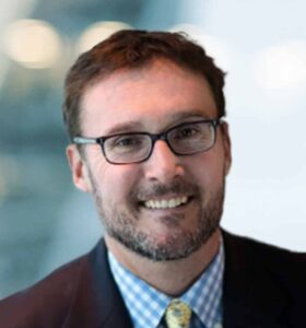 A man with short brown hair and a beard, wearing glasses, a dark suit, a blue checkered shirt, and a yellow tie, smiles at the camera against a blurred background.