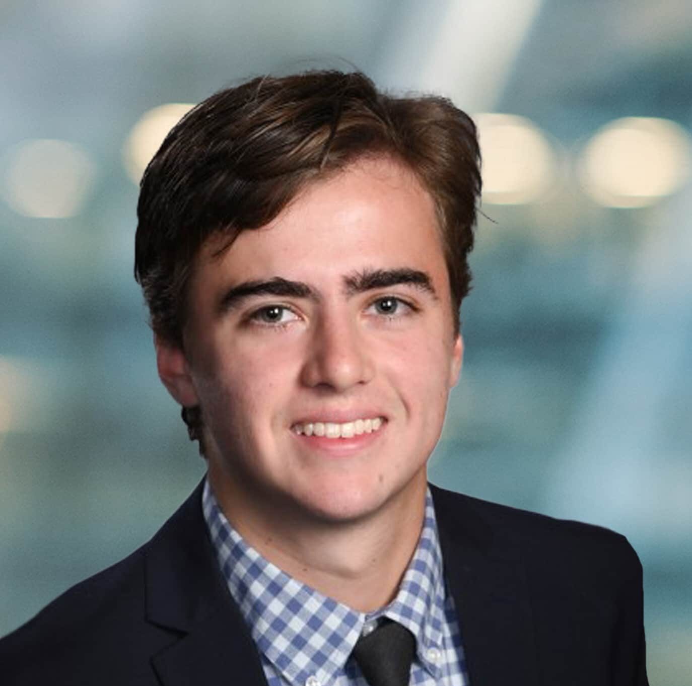 A young man with short brown hair wearing a dark blazer, checkered shirt, and tie, smiling in front of a blurred blue and white background.
