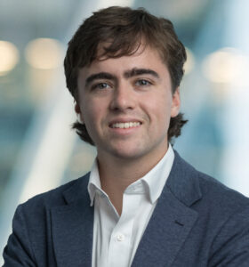 A young man with wavy brown hair, wearing a white shirt and blue blazer, smiles at the camera with arms crossed. The background is blurred with blue and white tones and soft lights.