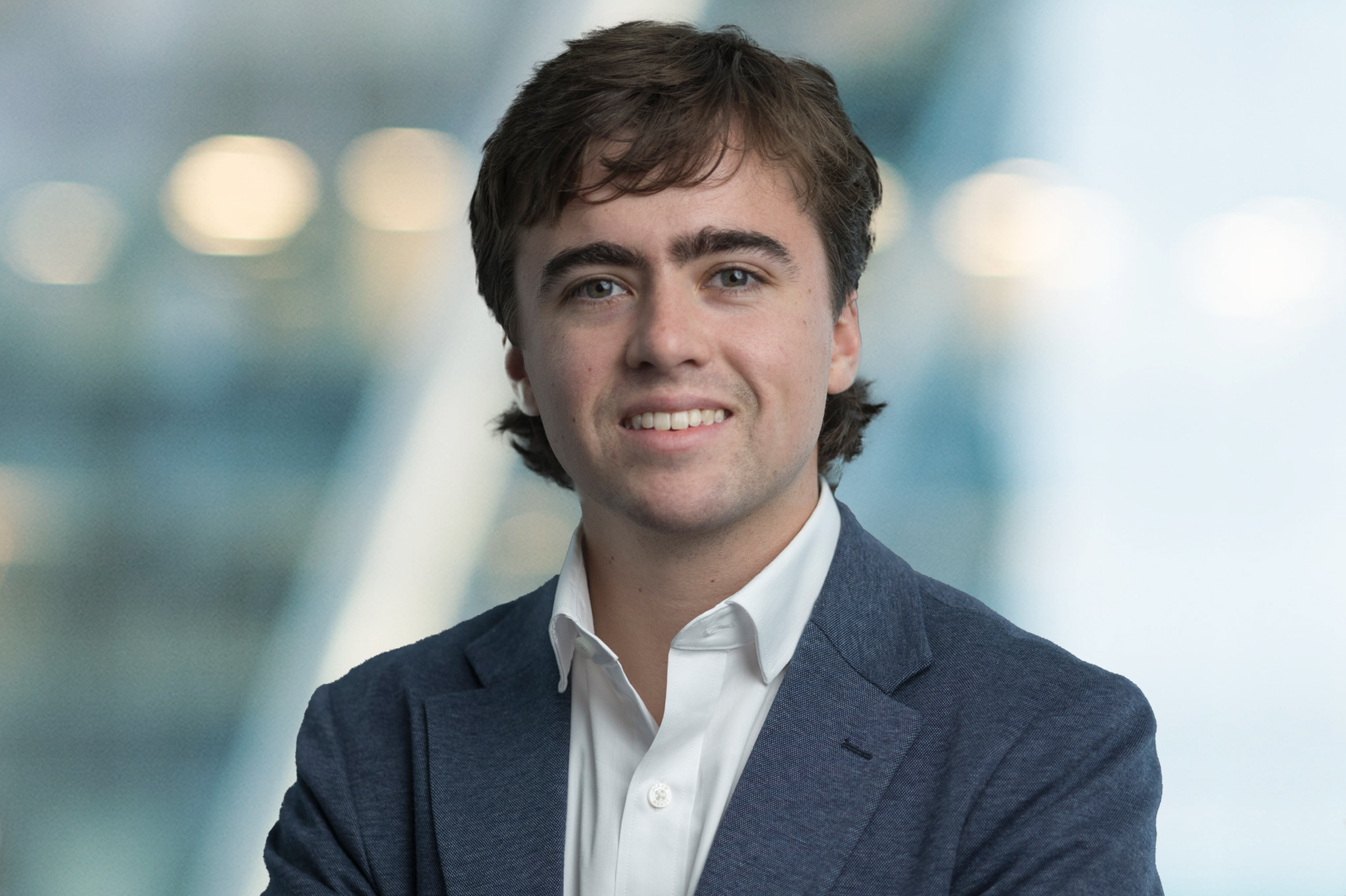 A young man with wavy brown hair, wearing a white shirt and blue blazer, smiles at the camera with arms crossed. The background is blurred with blue and white tones and soft lights.