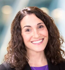 A woman with curly brown hair, wearing a black blazer and purple top, smiling at the camera. The background is softly blurred with blue and white tones.