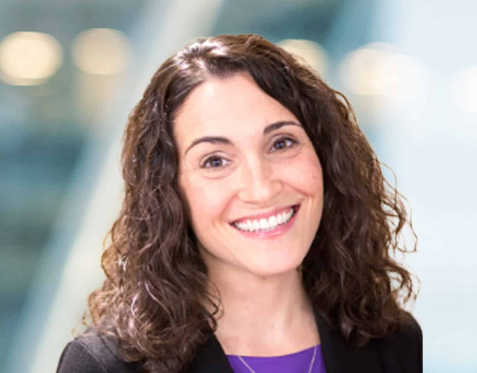 A woman with curly brown hair, wearing a black blazer and purple top, smiling at the camera. The background is softly blurred with blue and white tones.