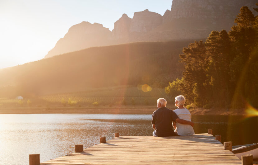 An elderly couple sits together on a wooden dock overlooking a calm lake, surrounded by trees and mountains, with warm sunlight illuminating the peaceful landscape.