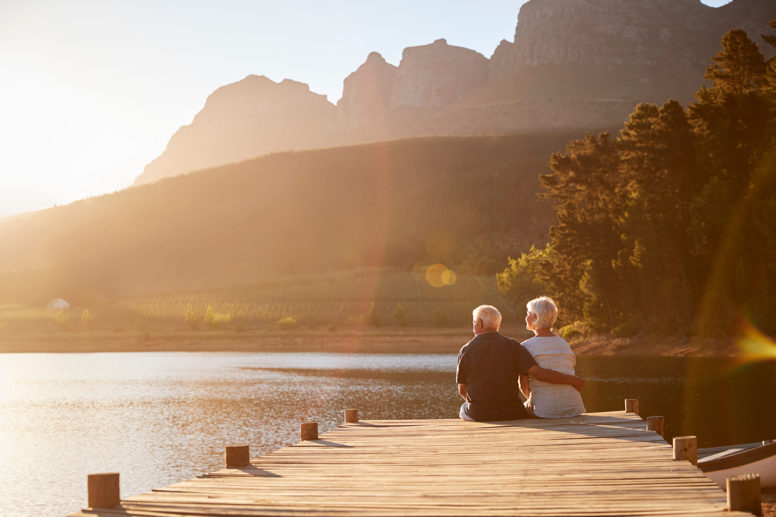 An elderly couple sits together on a wooden dock overlooking a calm lake, surrounded by trees and mountains, with warm sunlight illuminating the peaceful landscape.