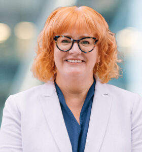 A person with curly, shoulder-length red hair and glasses, wearing a light lavender blazer over a dark blue top, smiles in front of a blurred background with soft lights.