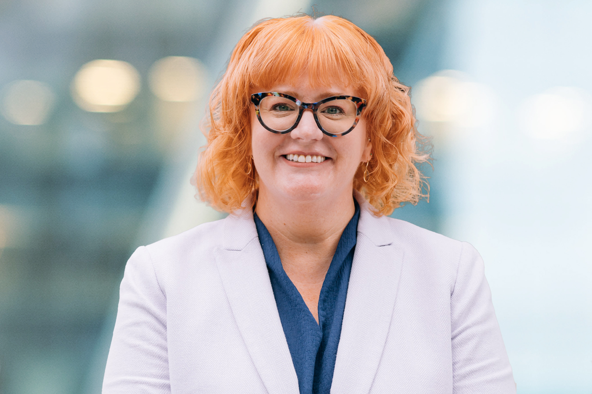 A person with curly, shoulder-length red hair and glasses, wearing a light lavender blazer over a dark blue top, smiles in front of a blurred background with soft lights.