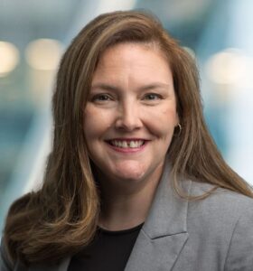 A woman with long brown hair wearing a gray blazer and black top smiles at the camera, with a blurred blue and white background.