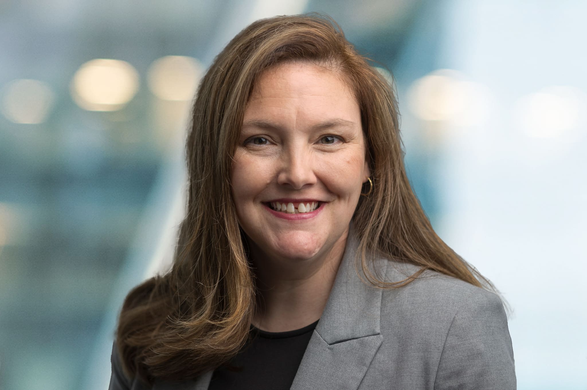 A woman with long brown hair wearing a gray blazer and black top smiles at the camera, with a blurred blue and white background.