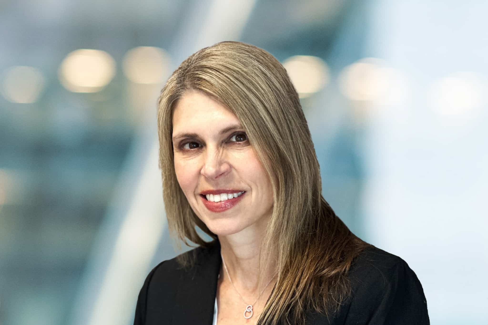 A woman with straight, light brown hair smiles at the camera. She is wearing a black blazer over a light top, with a necklace, and is posed against a softly blurred background with blue and white tones.