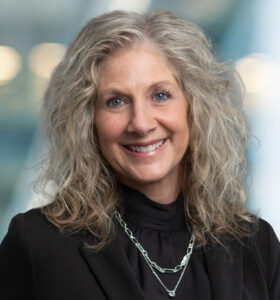 A middle-aged woman with curly gray hair, wearing a black blazer and layered silver necklaces, smiles in front of a softly blurred background with blue and yellow tones.
