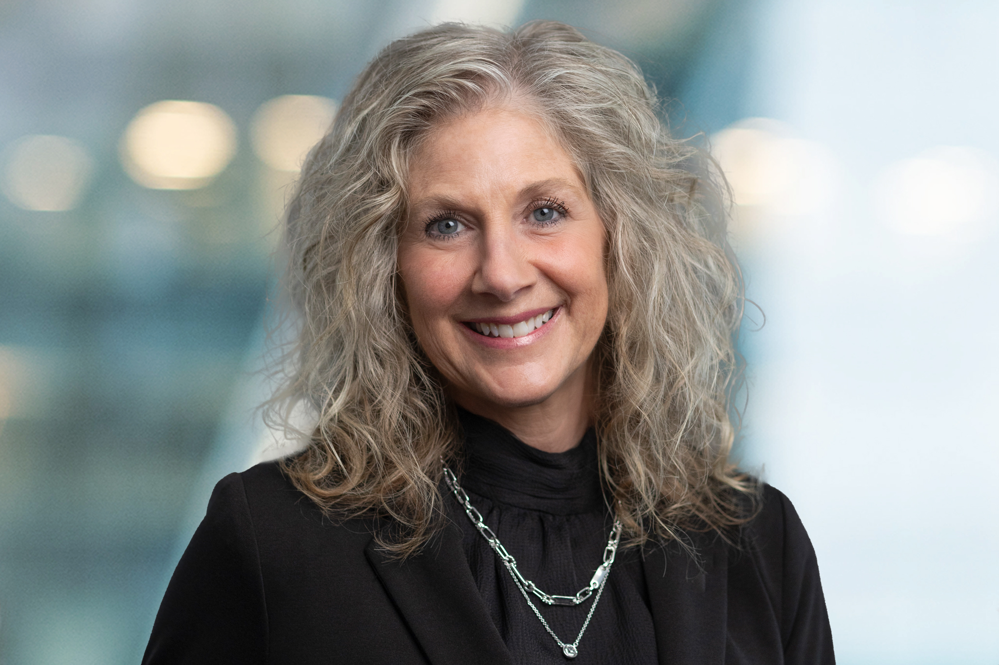 A middle-aged woman with curly gray hair, wearing a black blazer and layered silver necklaces, smiles in front of a softly blurred background with blue and yellow tones.