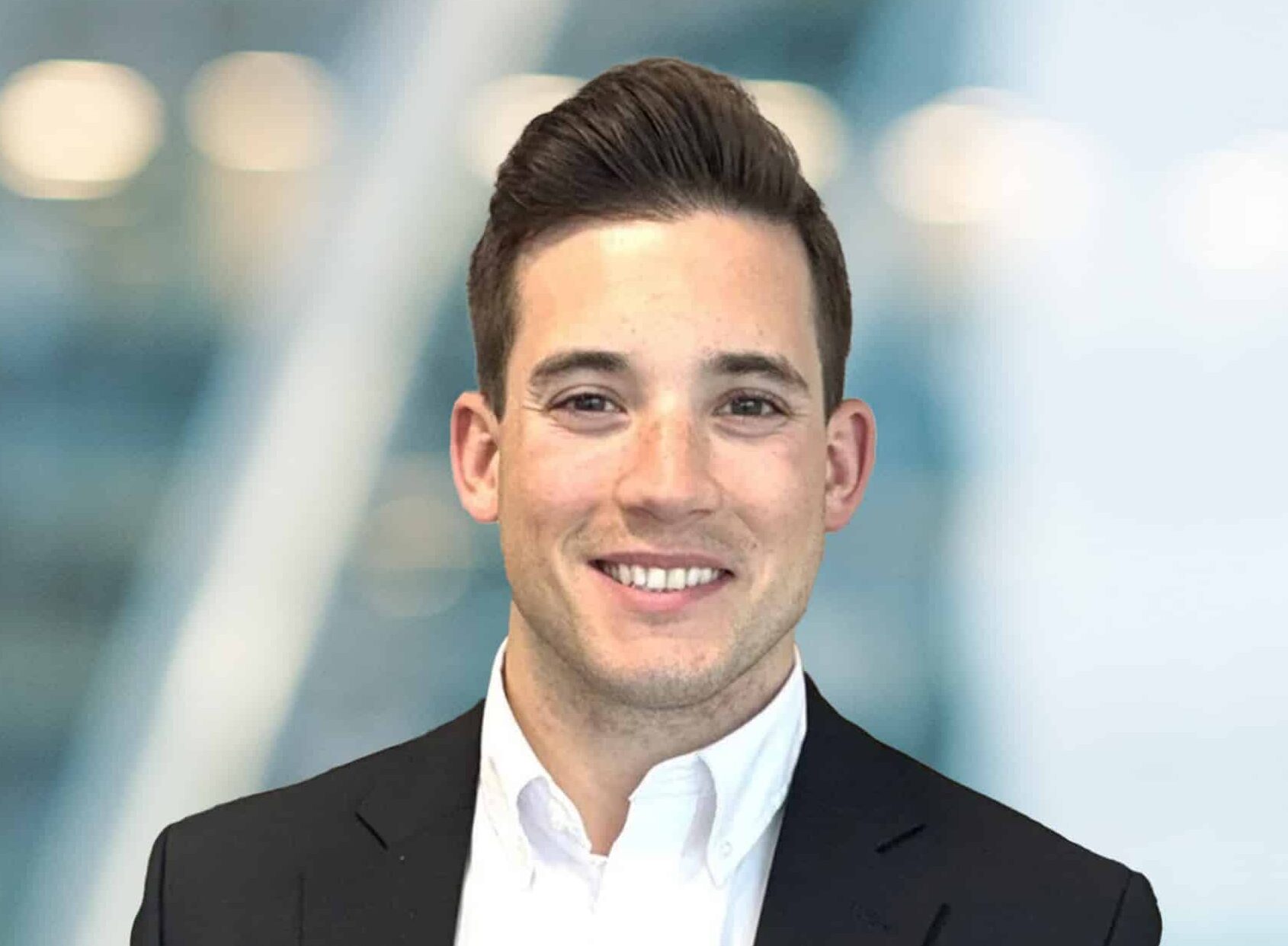 A young man in a dark suit and white dress shirt smiles at the camera. The background is softly blurred with blue and white tones, suggesting a professional setting.