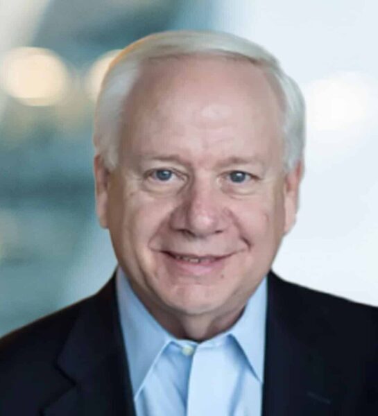 An older man with short white hair, wearing a dark suit jacket and a light blue collared shirt, smiles at the camera against a blurred background.