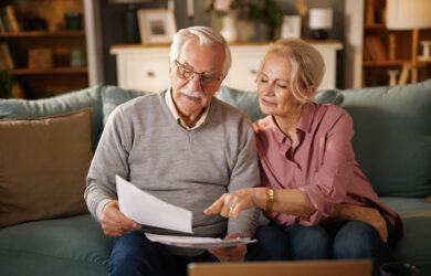 An older couple sits on a couch at home, looking at documents together. The man holds papers while the woman points at them, both appearing focused. A laptop is open on the table in front of them.