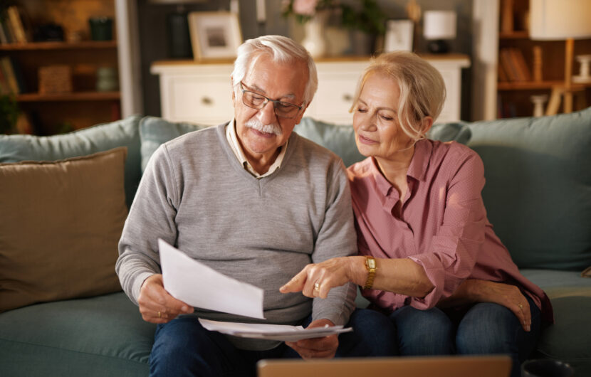 An older couple sits on a couch at home, looking at documents together. The man holds papers while the woman points at them, both appearing focused. A laptop is open on the table in front of them.