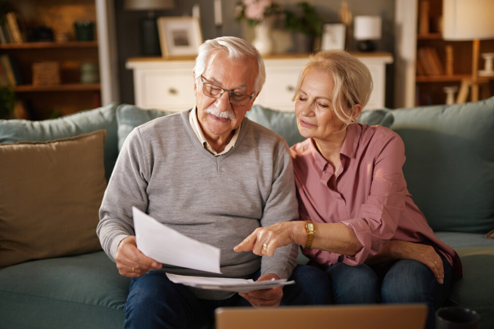 An older couple sits on a couch at home, looking at documents together. The man holds papers while the woman points at them, both appearing focused. A laptop is open on the table in front of them.