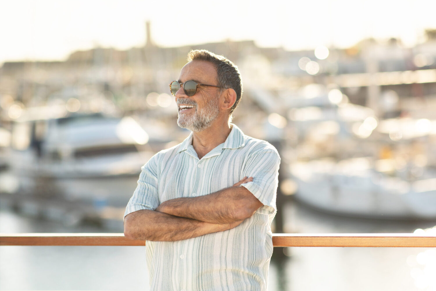 A smiling older man with gray hair and a beard stands with arms crossed, wearing sunglasses and a striped shirt, in front of a marina with blurred boats in the background on a sunny day.