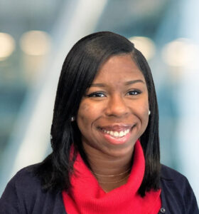 A woman with straight, shoulder-length black hair smiles at the camera. She is wearing a red turtleneck sweater and a dark jacket. The blurred background features cool tones and soft lighting.