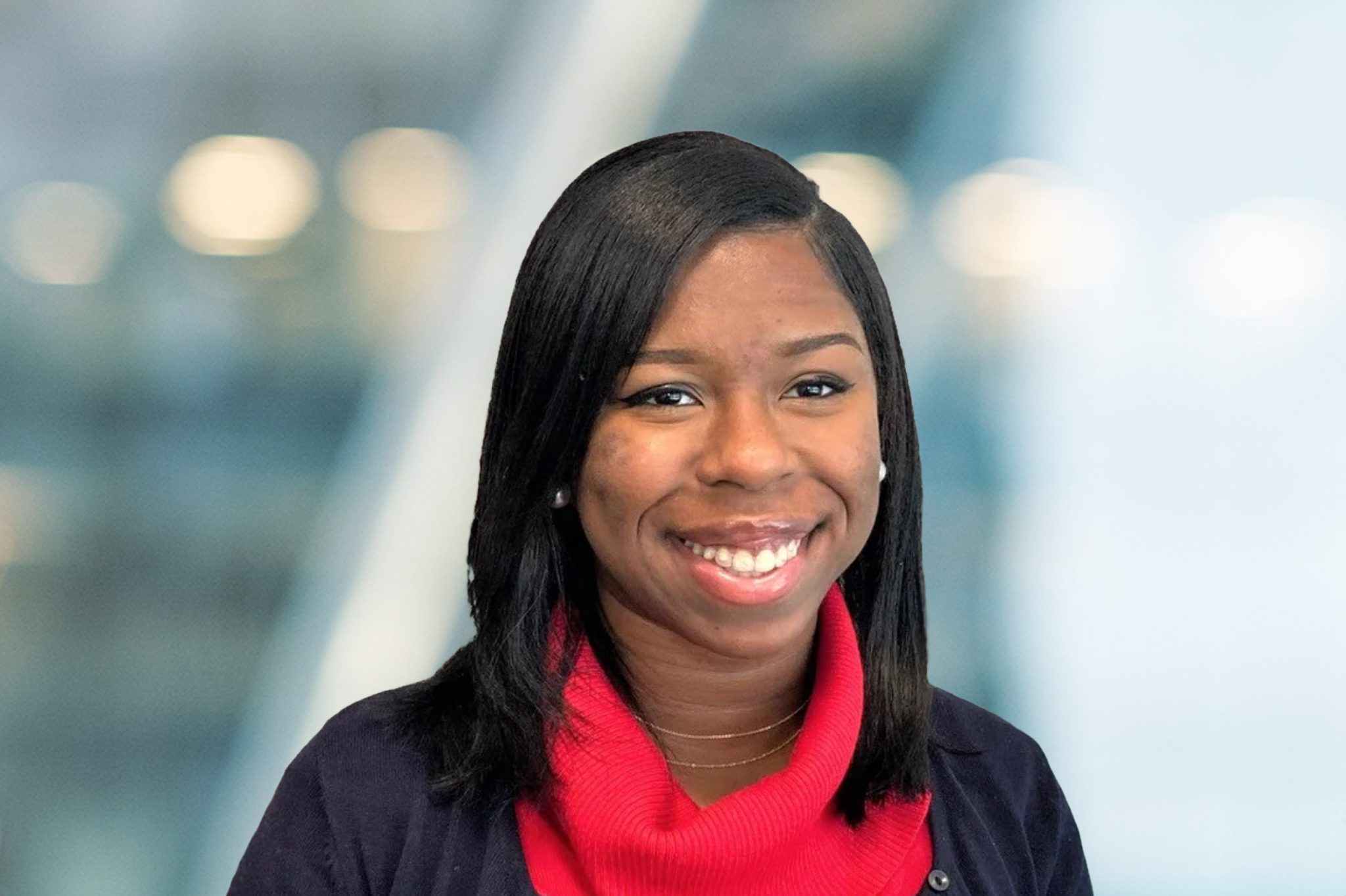 A woman with straight, shoulder-length black hair smiles at the camera. She is wearing a red turtleneck sweater and a dark jacket. The blurred background features cool tones and soft lighting.