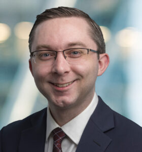 A smiling man wearing glasses, a dark suit, white shirt, and patterned tie, poses in front of a blurred, modern office background with soft lighting.