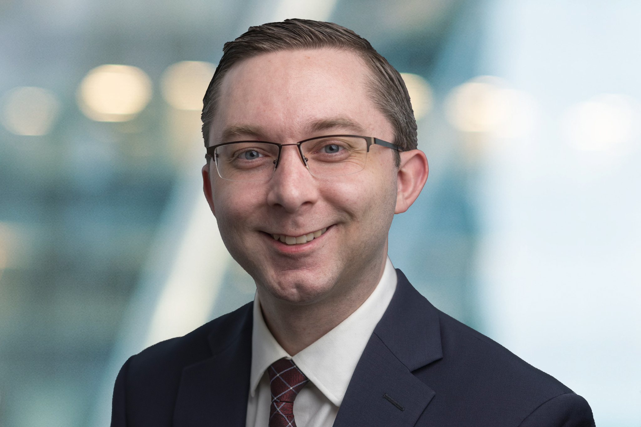 A smiling man wearing glasses, a dark suit, white shirt, and patterned tie, poses in front of a blurred, modern office background with soft lighting.