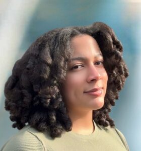 A person with curly, shoulder-length hair smiles softly while standing against a blurred, light blue background. They are wearing a light green ribbed top.