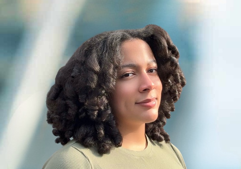 A person with curly, shoulder-length hair smiles softly while standing against a blurred, light blue background. They are wearing a light green ribbed top.